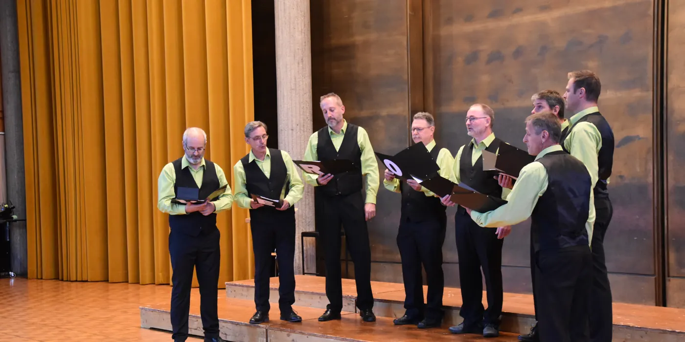 Les huit chanteurs de l’Octuor Veronika sur scène, chemises vertes et gilets noirs, devant un rideau doré