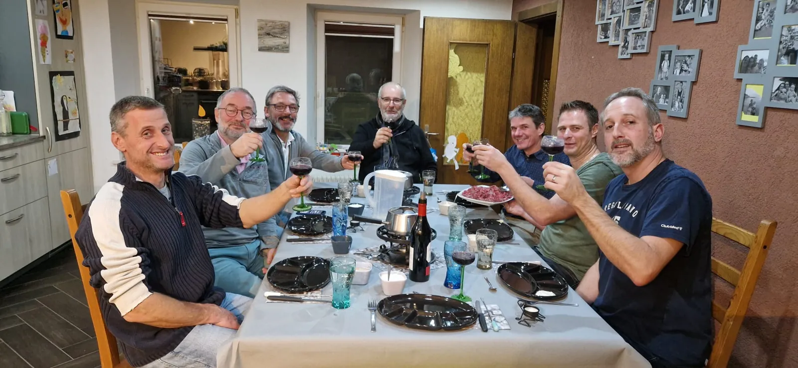 Sept des chanteurs attablés, verre levé, autour d’une fondue